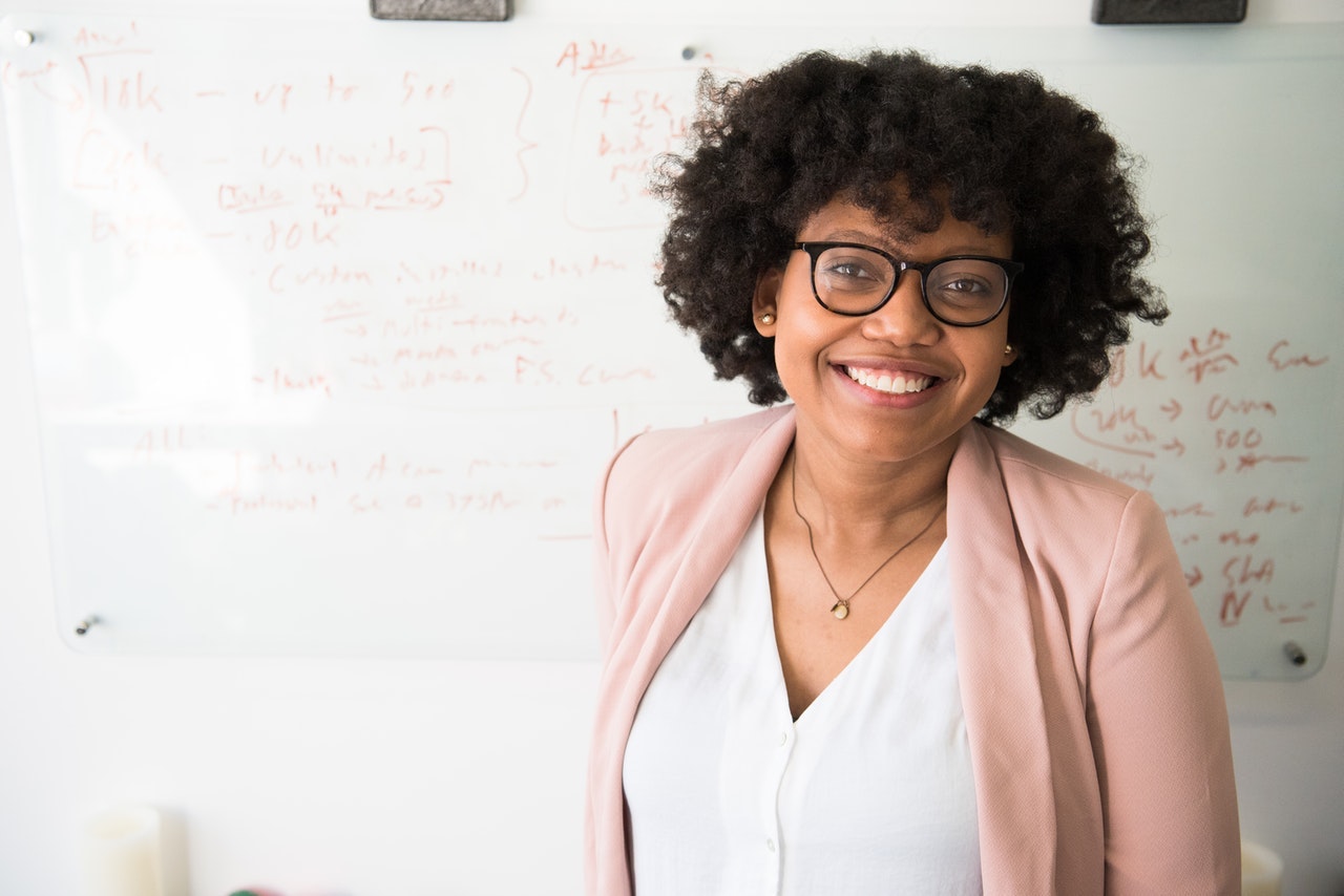 A teacher in a pink blazer stands in front of a whiteboard